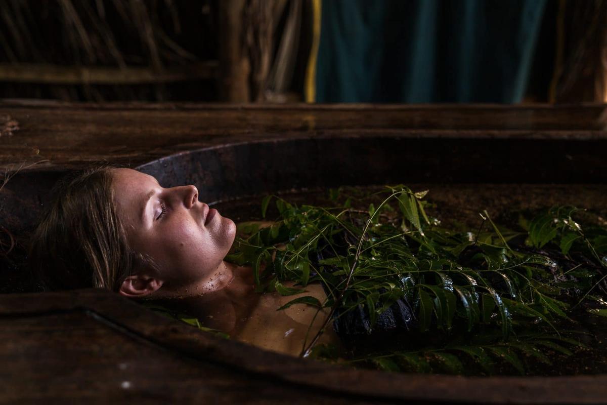 A guest resting with closed eyes in a carved wooden herbal bath, fresh neem branches floating on the dark herb-infused water under a thatched roof