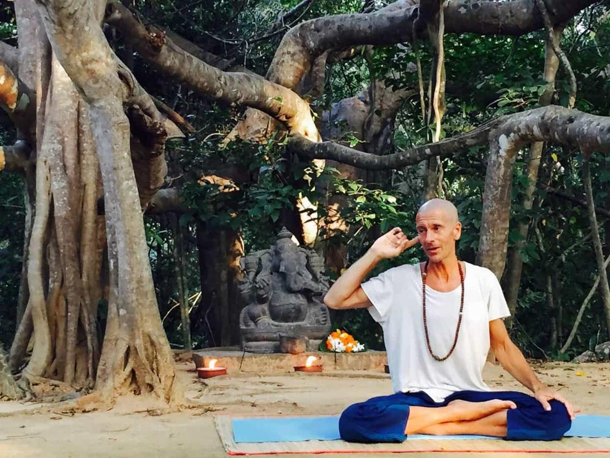 A shaven-headed yoga teacher in white sits cross-legged on a mat, gesturing mid-instruction beneath a huge banyan tree with a stone Ganesh statue, flowers and lit oil lamps behind him.