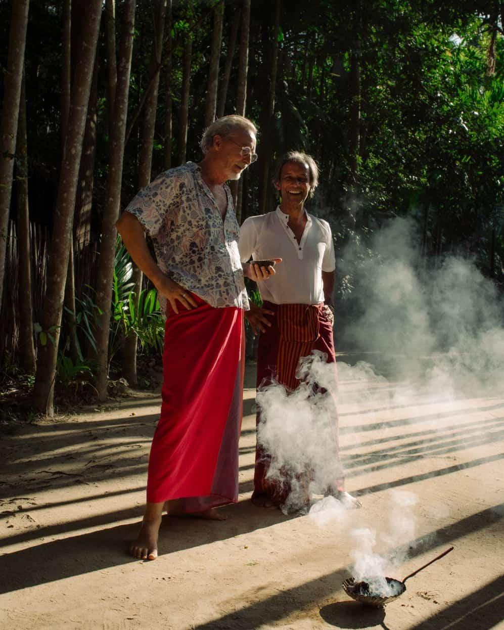Founders Giles Scott and Viren Perera laughing on a sandy path between tall trees at Ulpotha, incense smoke rising at their feet.