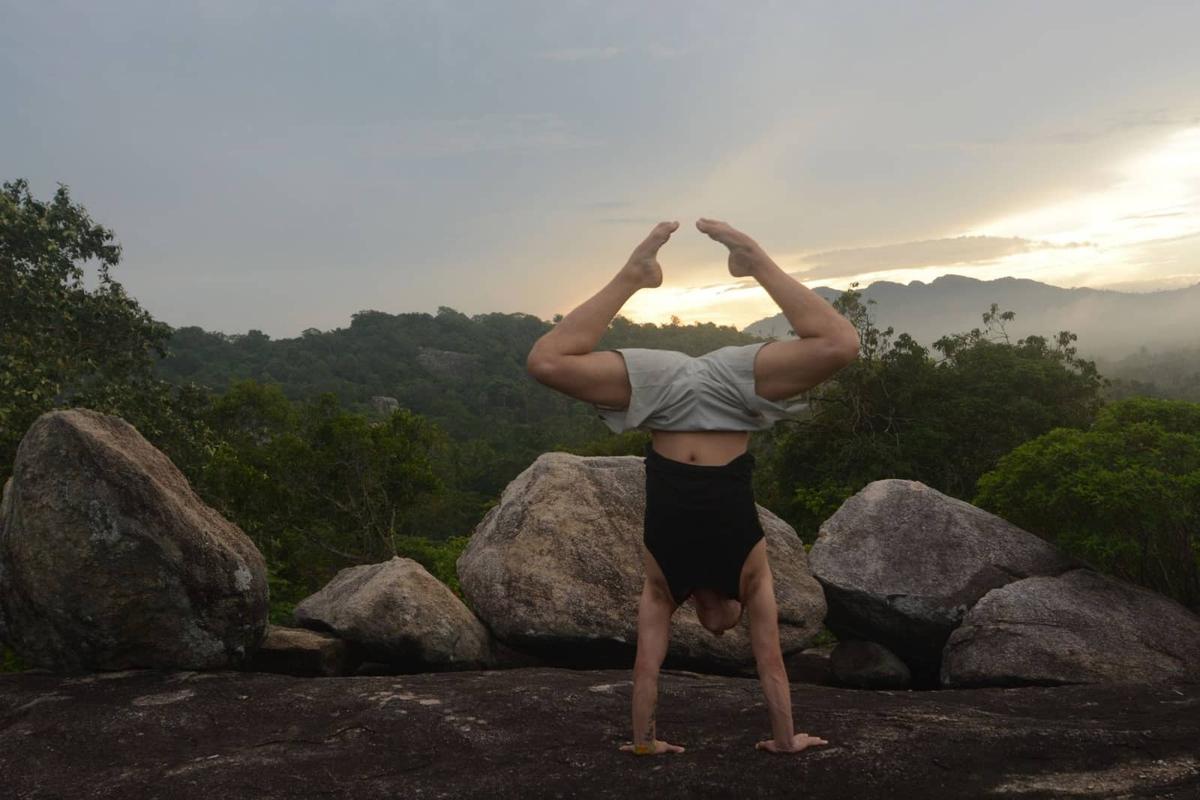 A man holding a straddle handstand on flat rock among giant boulders as the sun sets behind misty jungle ridges.
