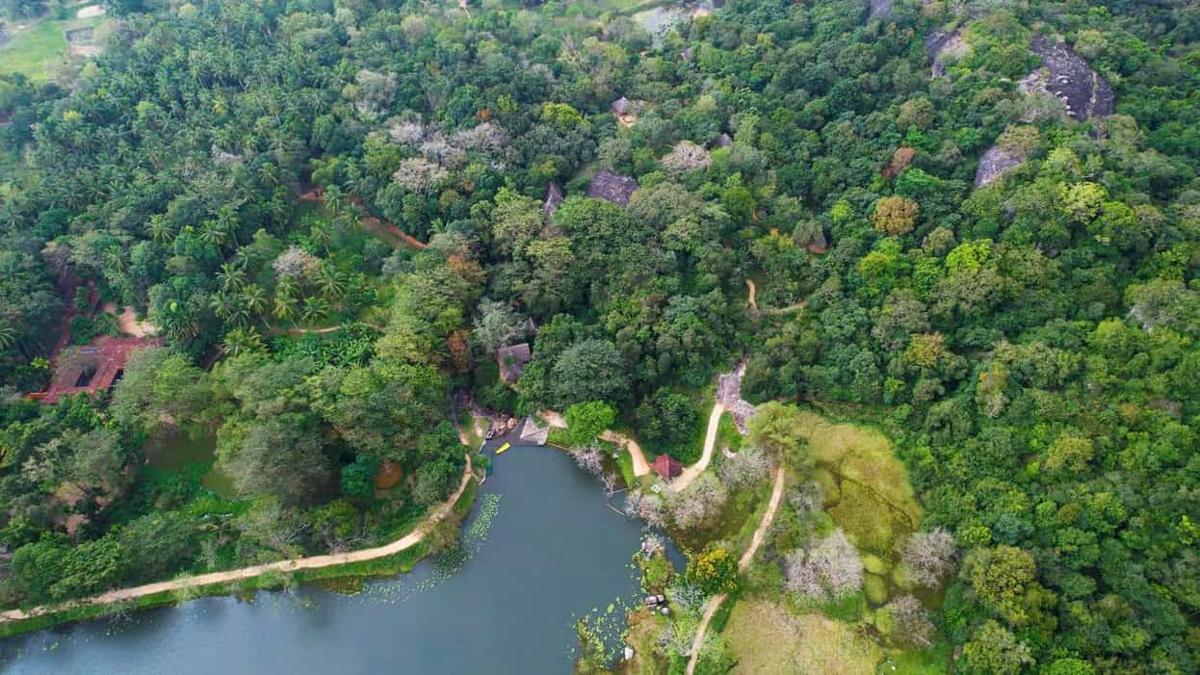 The lake edge from above: jungle canopy, granite boulders, a sandy path along the shore and thatched roofs hidden in the trees