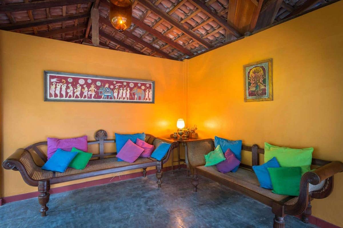 A marigold-yellow lounge under exposed roof beams, with antique cane settees piled with jewel-tone cushions and a brass lamp aglow
