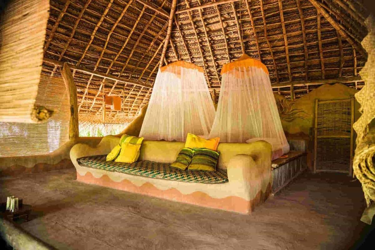 Inside a wattle-and-daub hut at night: a sculpted platform bed beneath two coned white mosquito nets hanging from the woven-palm thatch