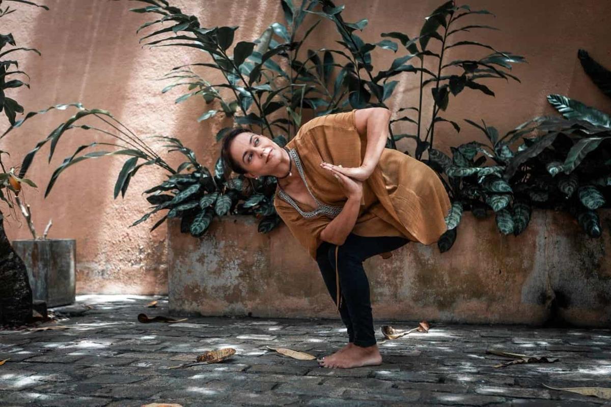 A woman in a mustard cover-up in a standing side-bend on mossy stone paving, before a salmon-pink wall lined with dark tropical plants.