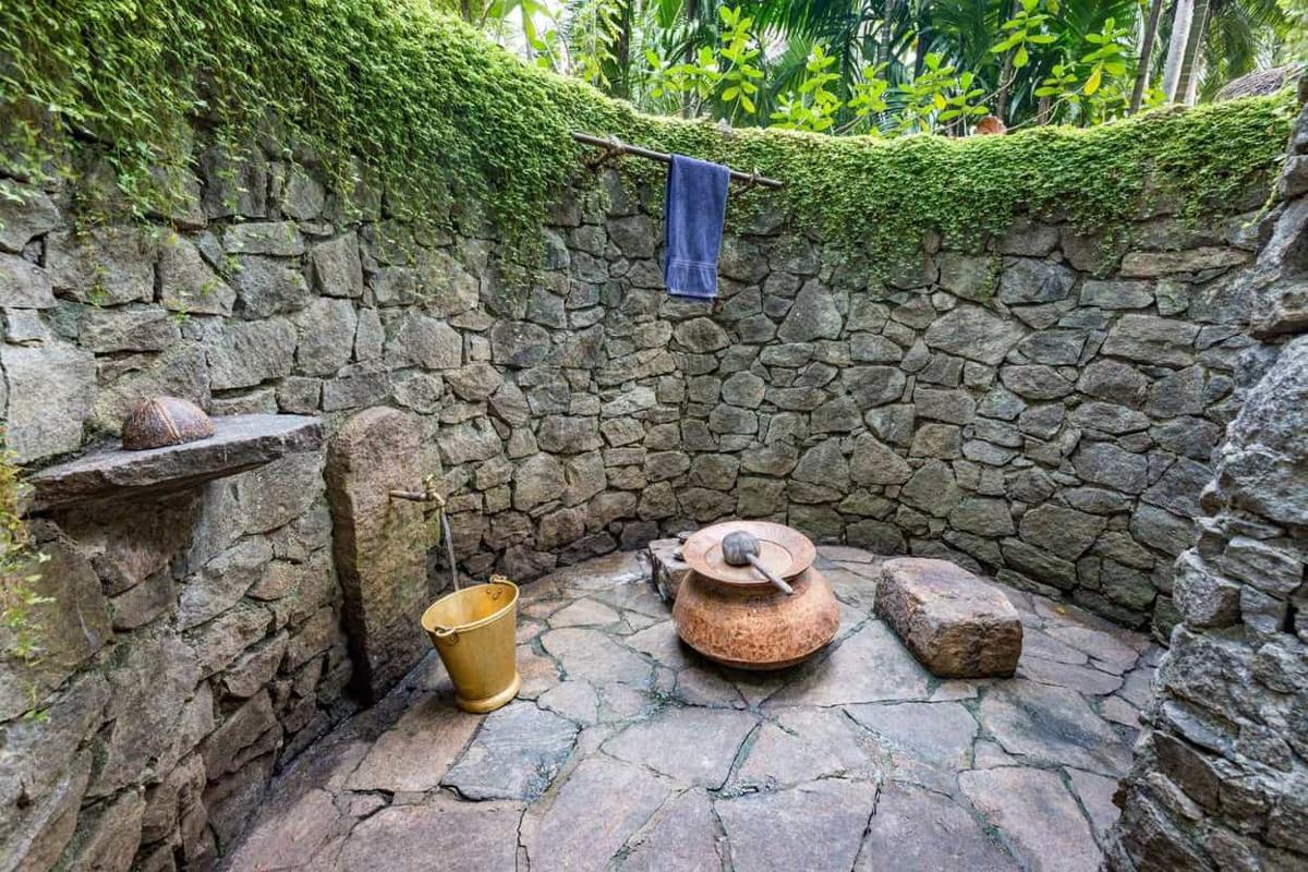 Open-air stone bathing enclosure with moss-covered walls, a brass bucket beneath a tap, a copper water pot on a flagstone floor, and jungle foliage above.