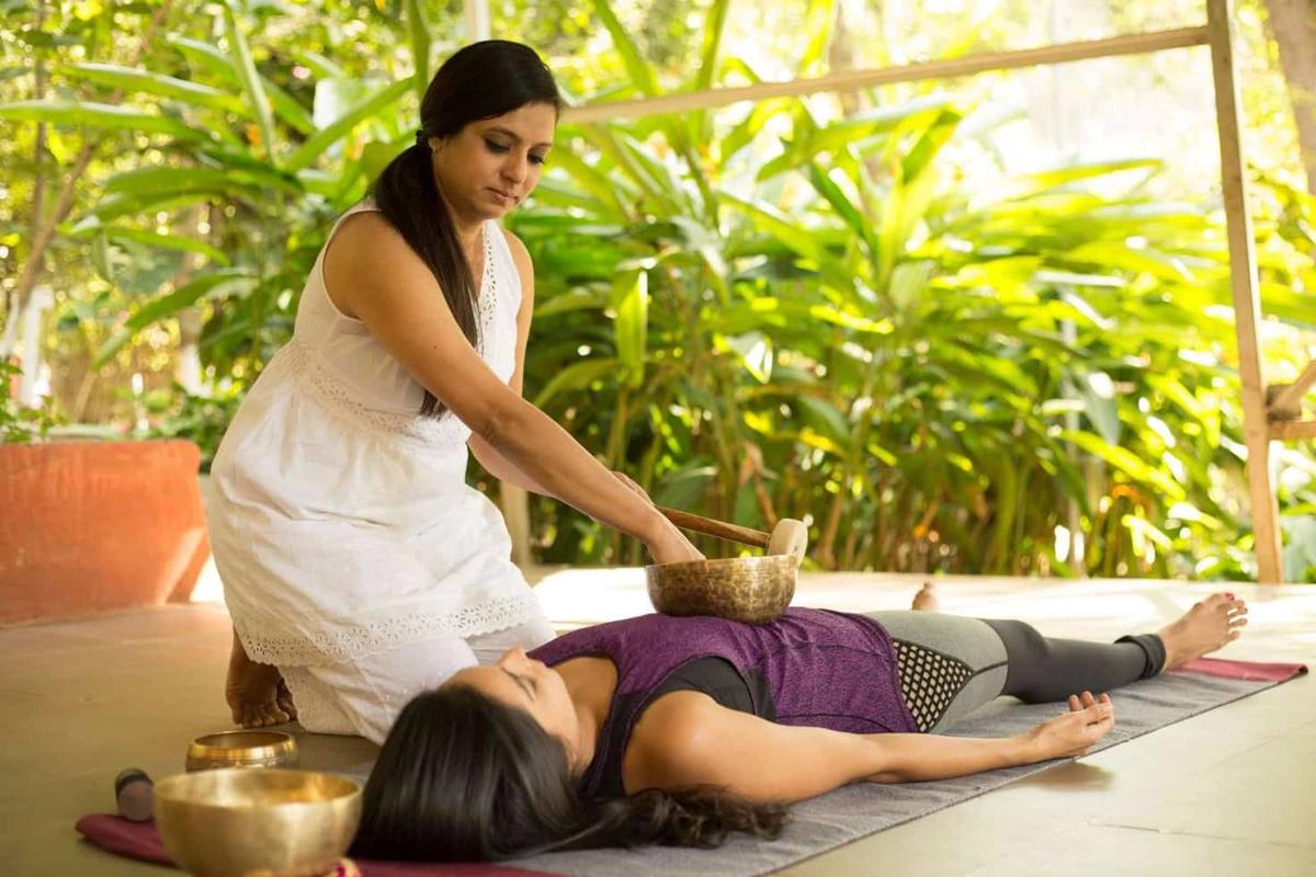 A therapist in white strikes a bronze singing bowl resting on the chest of a guest lying in an open pavilion, lush foliage behind.