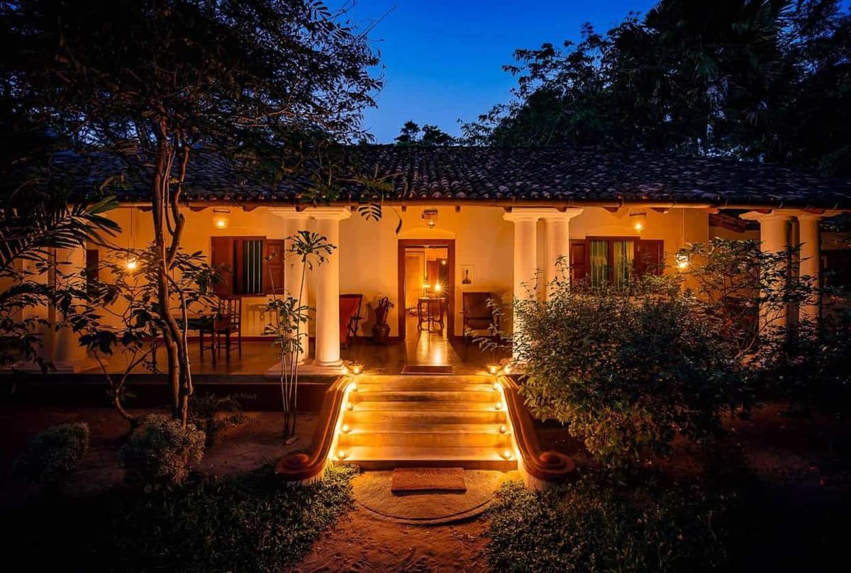 The white-columned veranda of the walawwa at deep-blue dusk, every step of the entrance stair lined with glowing candle lanterns