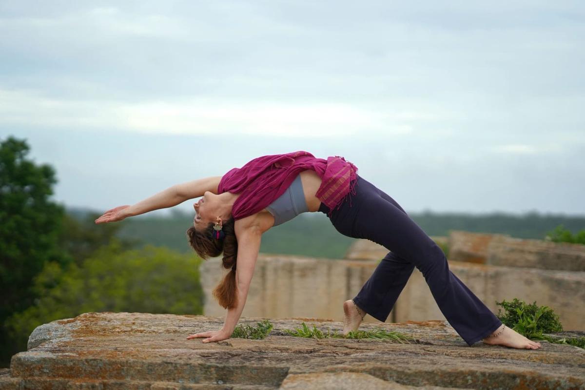 A woman in a magenta scarf arching into wild thing pose on a lichen-covered granite outcrop, the jungle canopy soft below.