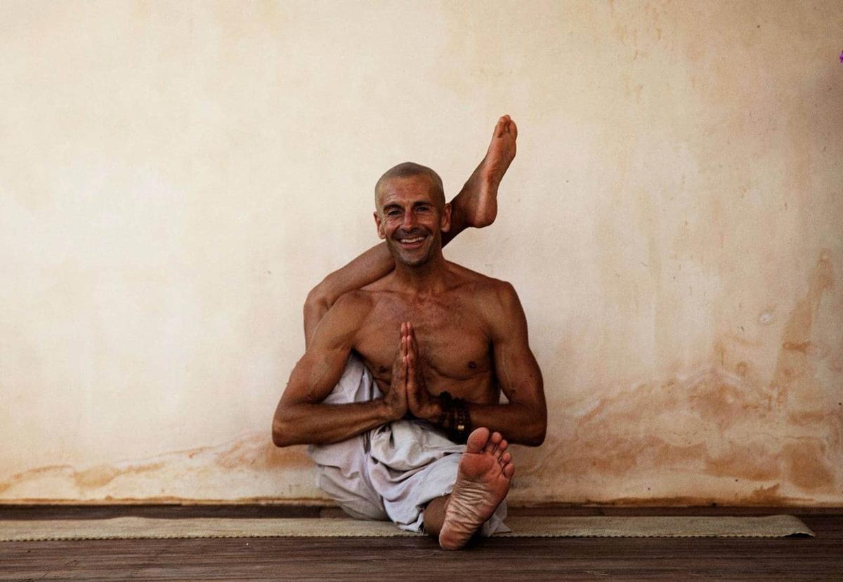 A shaven-headed yogi seated against a weathered lime-plaster wall, one leg behind his head, hands in namaste, beaming.