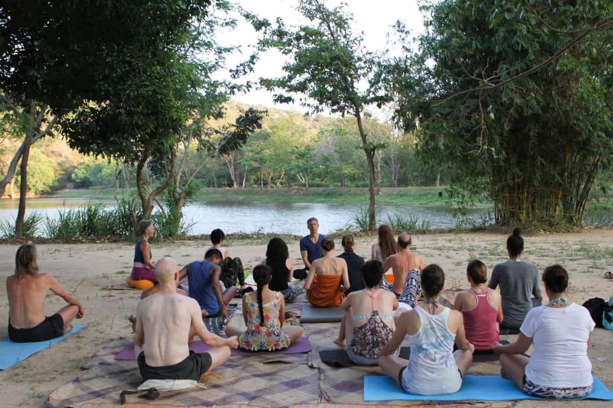 A class seated on mats on the sand by the lake, under the trees