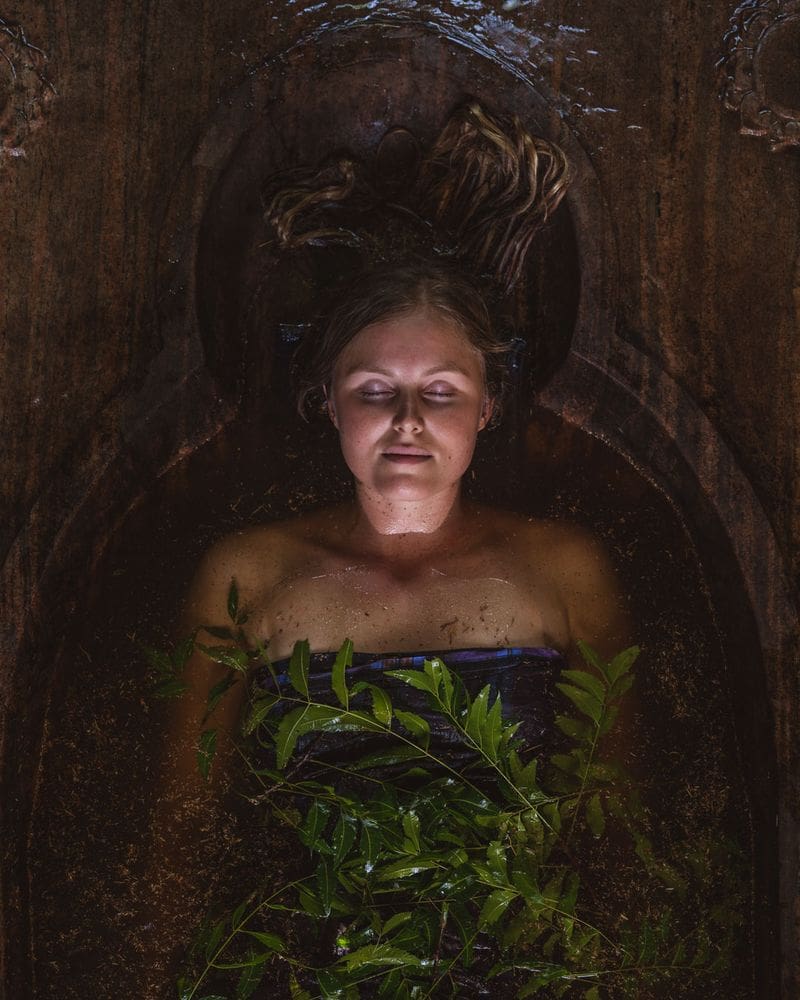 Seen from above, a guest lies with closed eyes in a carved wooden tub, neem branches spread across the herb-infused water