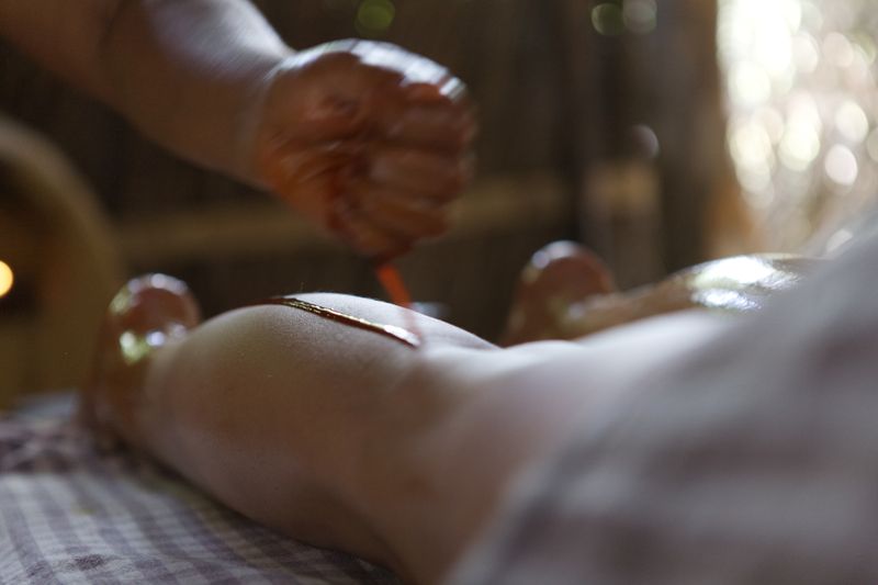 A therapist's hand wrings warm herbal oil from a cloth in a thin stream onto a guest's back, the treatment room soft-focus behind