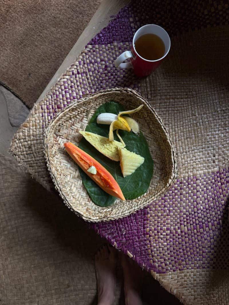 A woven basket with papaya, banana and cake arranged on a green leaf, next to a red mug of tea on handwoven mats.