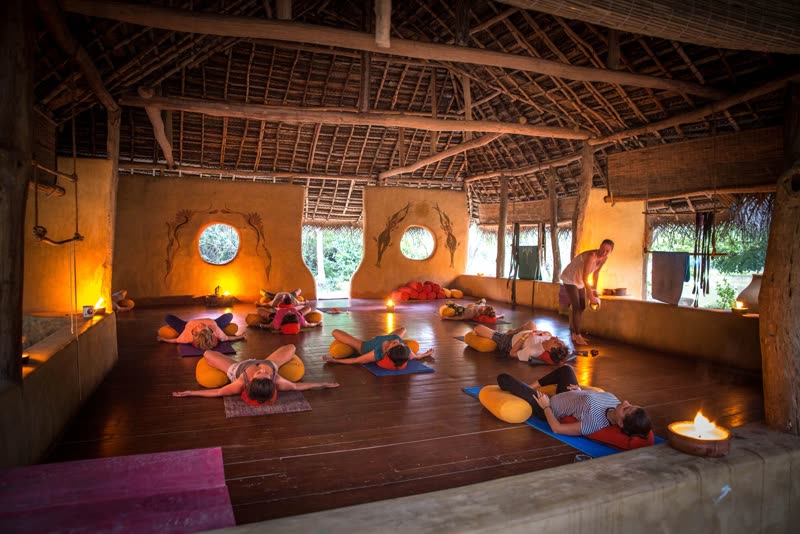 Guests lying over yellow bolsters on yoga mats in a candlelit open-air shala with thatched roof and ochre walls at dusk.