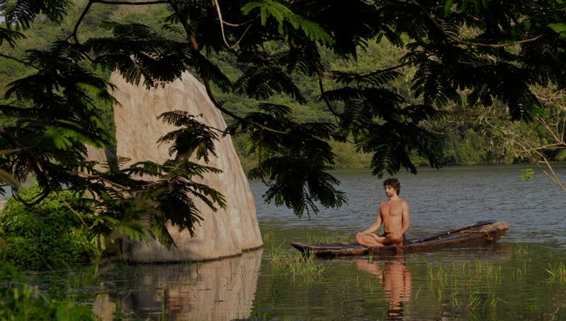 A man meditates cross-legged on a wooden dugout canoe on a calm lake, beside a tall granite rock and jungle foliage.