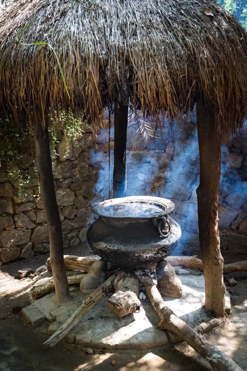 Large blackened cauldron steaming over an open wood fire under a thatched canopy, with smoke rising against a stone wall.