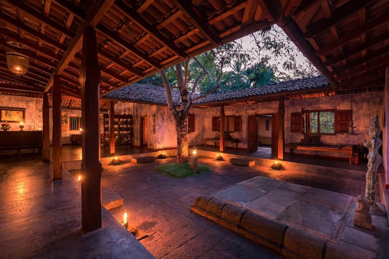 The stone-paved open-roof courtyard at twilight, a tree growing through it, dozens of small candles and lanterns glowing warm against the blue evening light