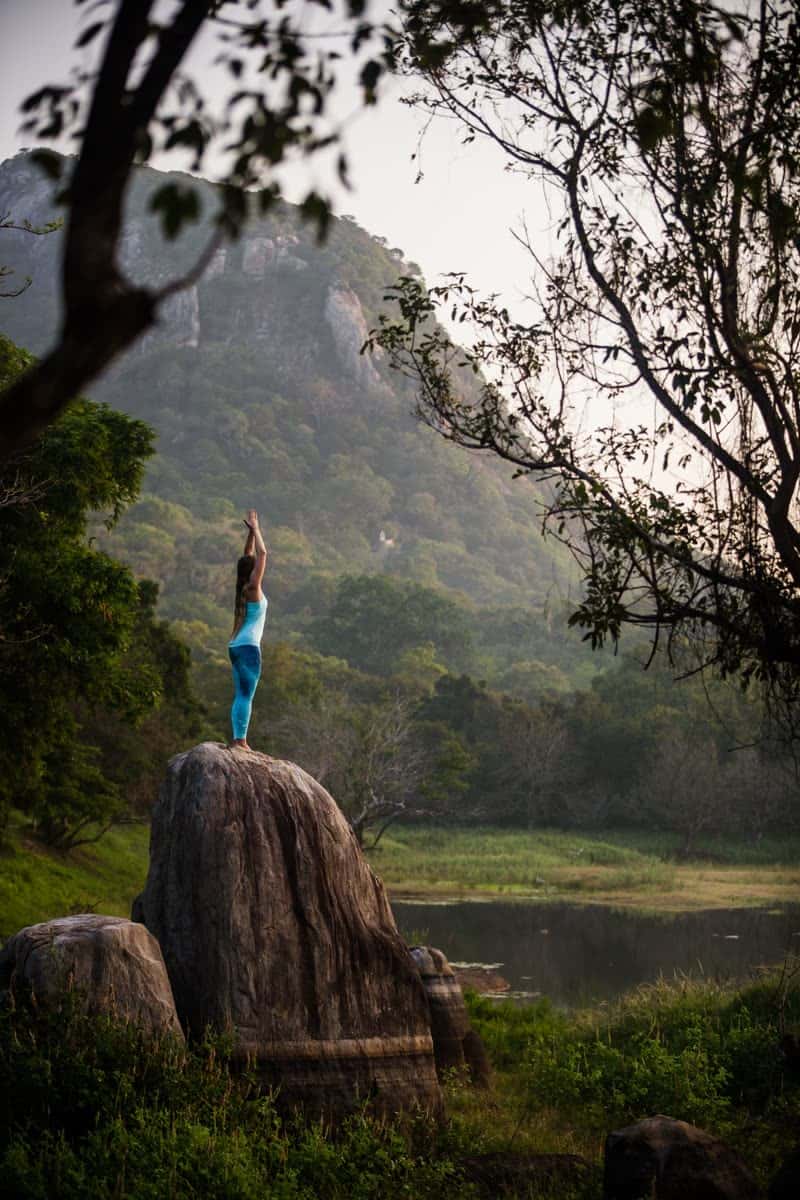 Woman in blue yoga clothes standing on a large boulder with arms stretched overhead, facing a misty forested mountain beside a lake at dawn.