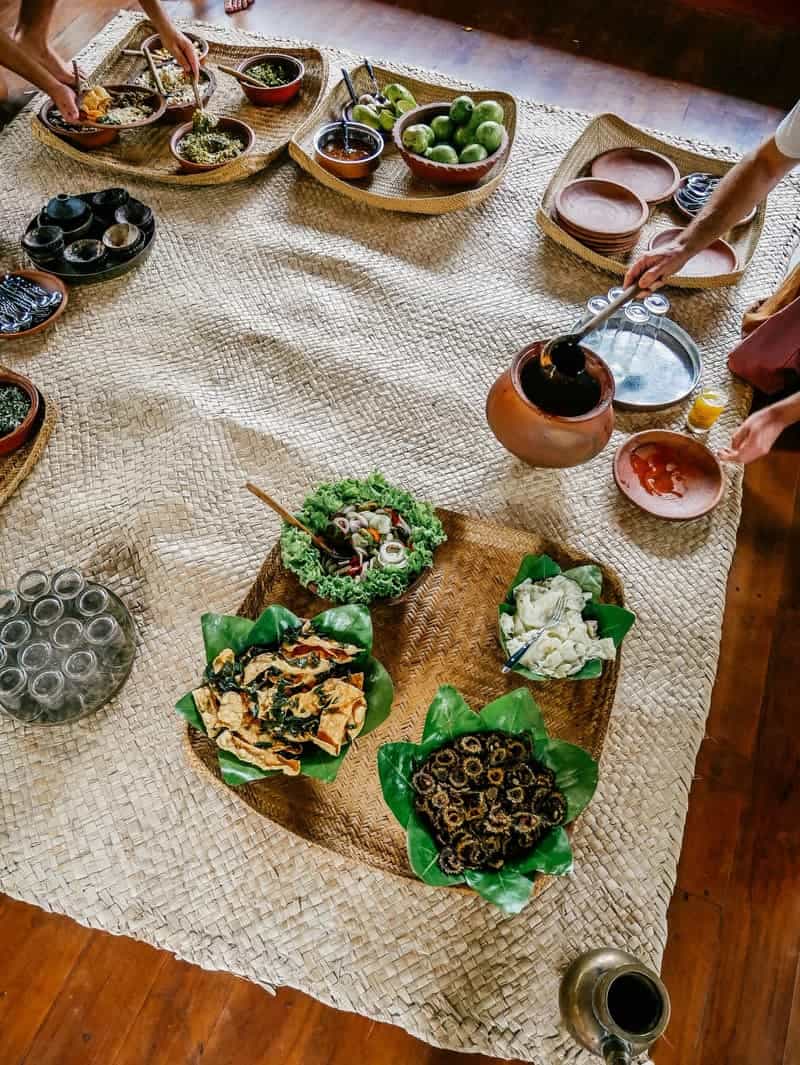 Overhead view of a communal floor dinner: dishes served on banana leaves in woven rattan trays laid out on rush mats, hands reaching in to serve