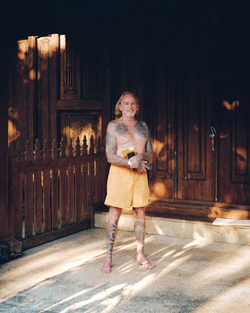 A weathered, heavily tattooed older man in orange shorts standing barefoot with a book in front of magnificent carved antique wooden doors, in dappled light