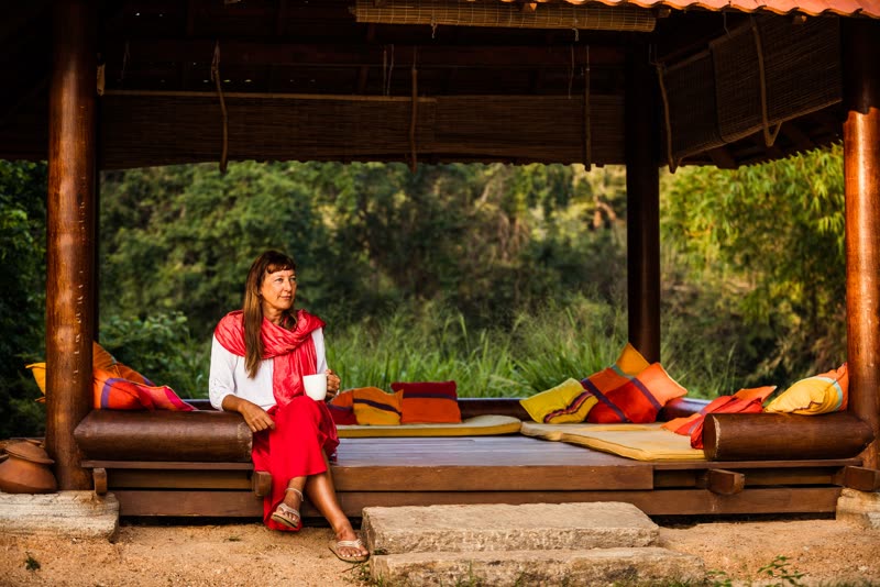 A woman in red sits with a mug of tea on the edge of an open-sided cushioned pavilion, with dense green jungle behind her.