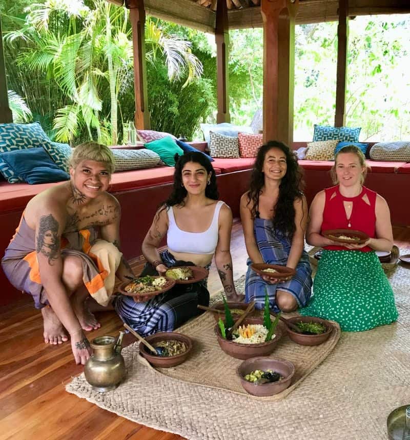 Four women guests sitting on woven mats around clay bowls of curry and rice in an open-air pavilion surrounded by jungle.