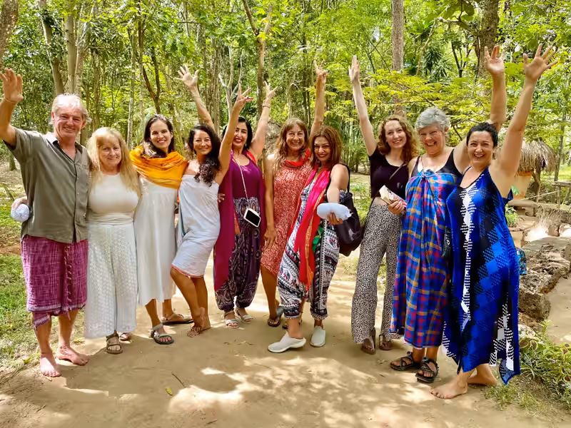 Ten happy retreat guests in bright sarongs raise their arms together on a sunlit sandy path beneath jungle trees.