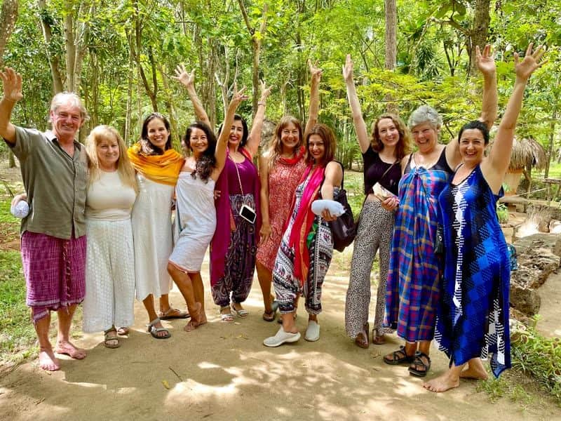 Ten happy retreat guests in bright sarongs raise their arms together on a sunlit sandy path beneath jungle trees.