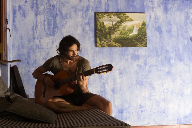 A man playing an acoustic guitar while seated on a striped daybed against a blue-washed wall with a jungle painting.