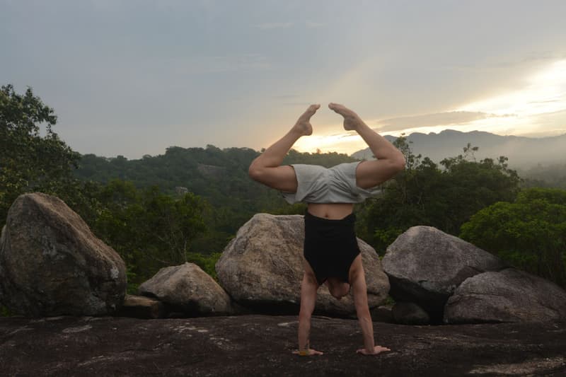 A man holds a straddle handstand on flat rock among giant boulders as the sun sets behind misty jungle ridges