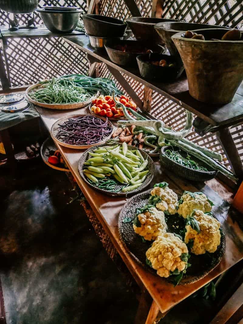 Baskets of fresh vegetables including beans, chillies, tomatoes and cauliflower arranged on a wooden table in an open-air kitchen with lattice walls.