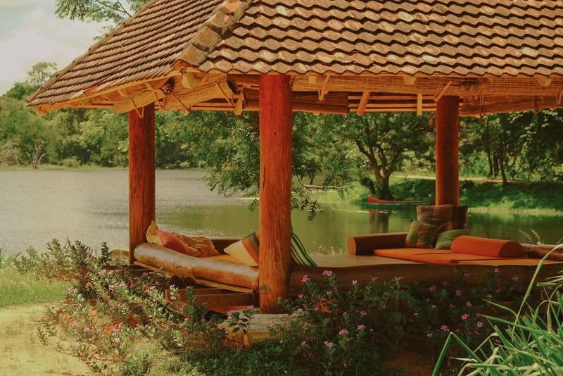 Open-sided pavilion with wooden daybeds and orange cushions overlooking a calm lake, with a red canoe moored among trees beyond.