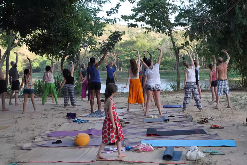A group yoga class stretching with raised arms on mats under trees beside the lake, a young girl walking among them.