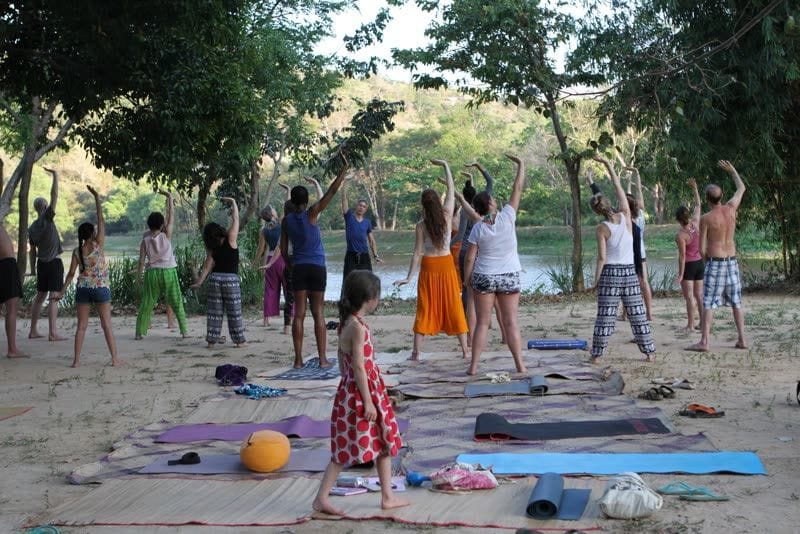 A group yoga class stretching with raised arms on mats under trees beside the lake, a young girl walking among them.