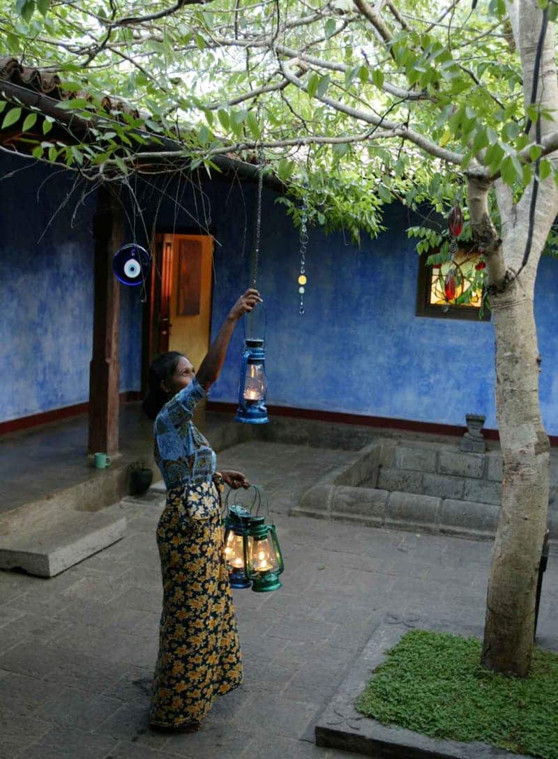 A Sri Lankan woman in a batik sarong reaching up to hang a lit blue-glass oil lantern from a tree in the indigo-walled courtyard at dusk, two green lanterns in her other hand