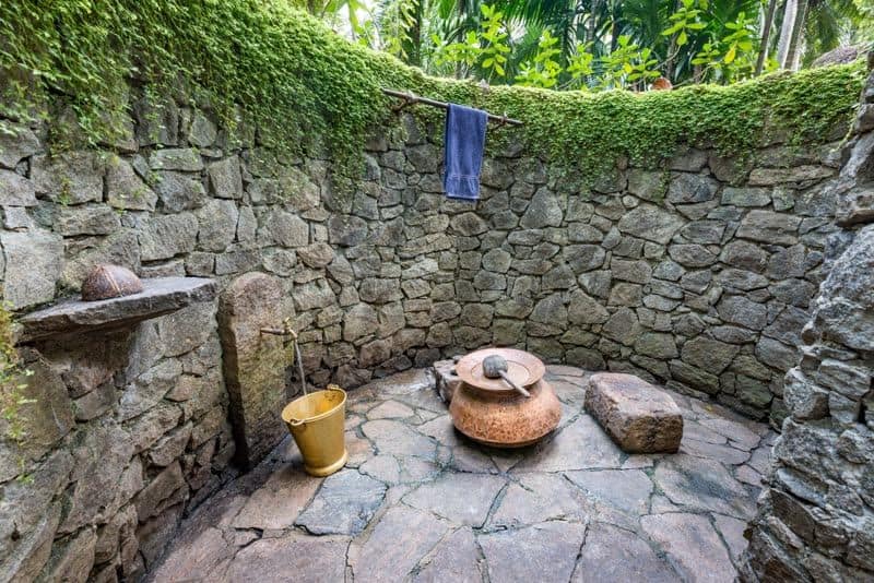 Open-air stone bathing enclosure with moss-covered walls, a brass bucket beneath a tap, a copper water pot on a flagstone floor, and jungle foliage above.
