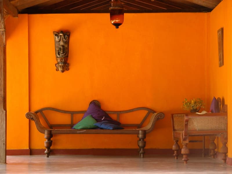 A vivid saffron-orange wall with an antique carved wooden daybed piled with silk cushions, a brass cobra mask and an amber glass lamp