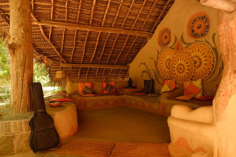 Open-air thatched lounge with earthen benches, bright cushions, a painted owl mural on the mud wall, and a guitar case leaning near the entrance.
