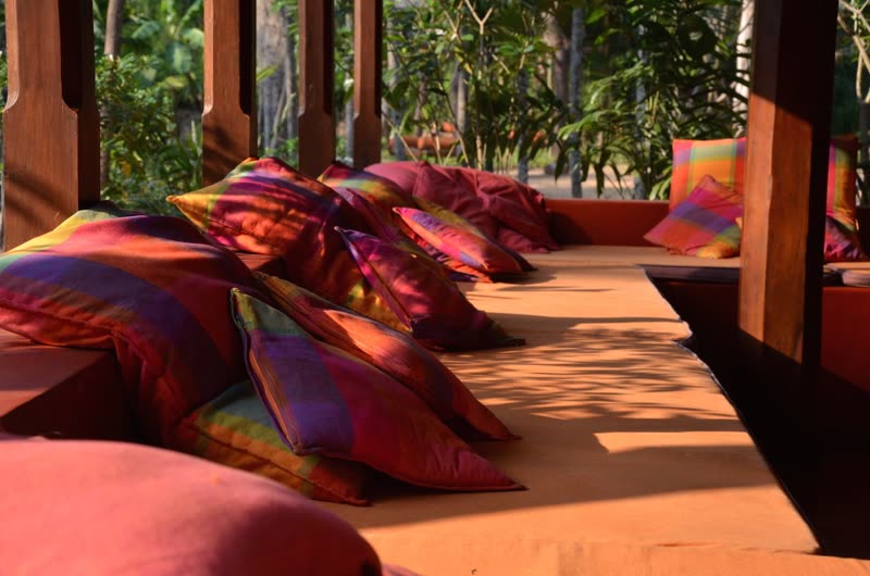 Colourful plaid cushions piled along a terracotta bench in an open wooden pavilion, with jungle foliage behind.
