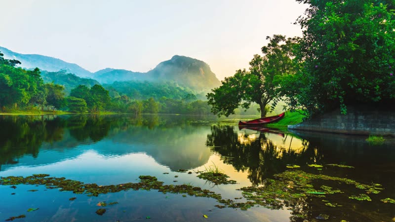 A red wooden canoe beached at the edge of a calm lake at dawn, with misty jungle-covered hills and a rock outcrop reflected in the still water.