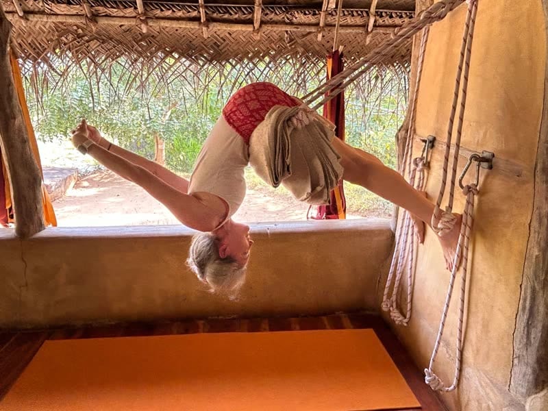 Woman hanging upside down in a rope-supported backbend against a yoga wall in an open-air thatched shala, jungle visible behind her.