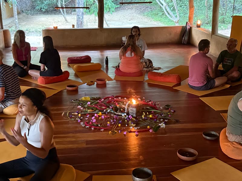 Retreat guests seated in a circle in an open-sided yoga shala around a flower mandala with a burning candle, with jungle greenery beyond.