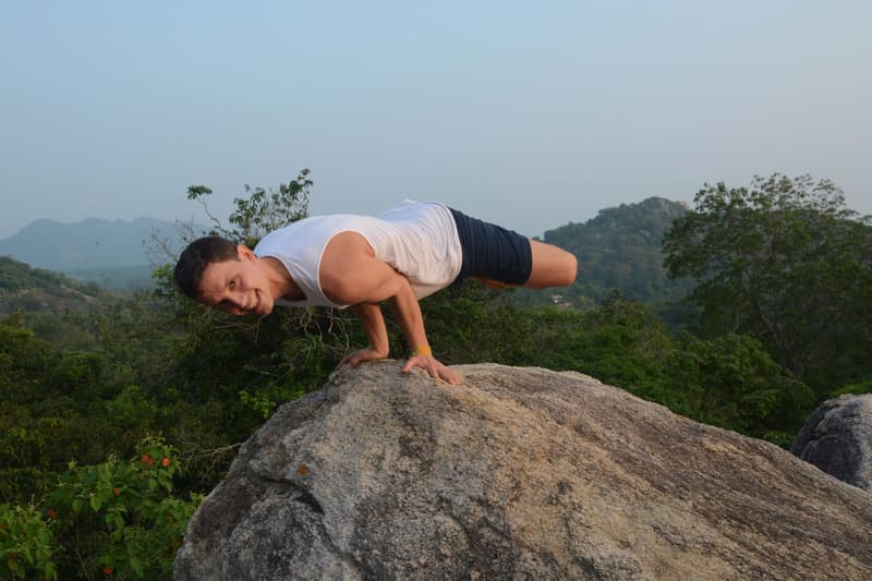 A young man balancing in side-crow arm balance on a granite boulder high above the jungle canopy, hazy hills beyond, grinning at the camera