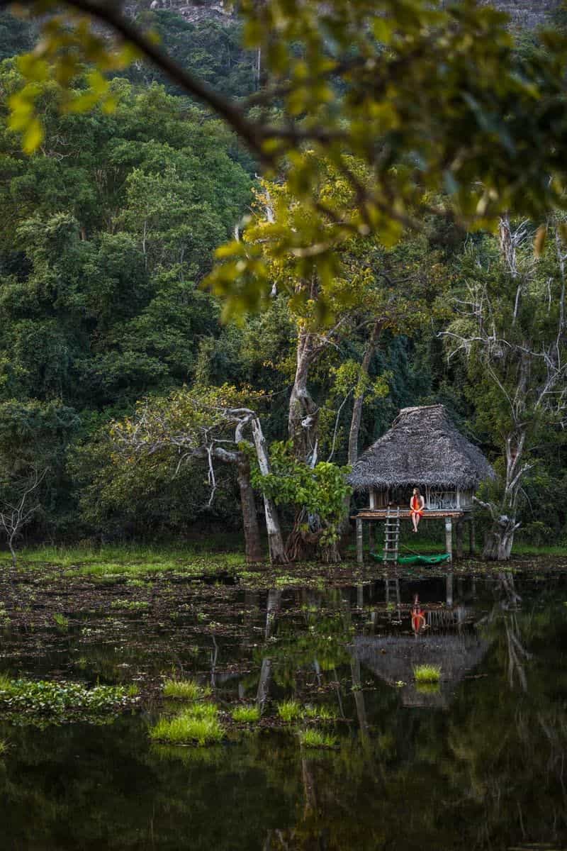 Woman seated on a stilted thatched pavilion over a calm jungle lake, her reflection in the water and a canoe moored beneath.