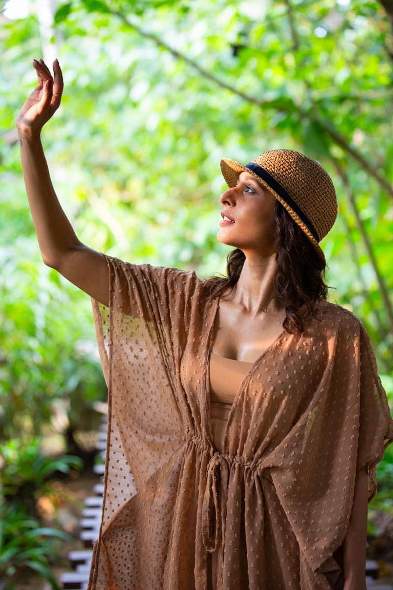A woman in a crochet straw hat and sheer caramel kaftan reaching up toward sunlit leaves on a green garden path