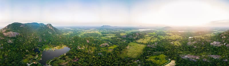 Wide aerial panorama of a green valley in Sri Lanka with a small lake, dense jungle, rice paddies and rocky hills fading into haze.