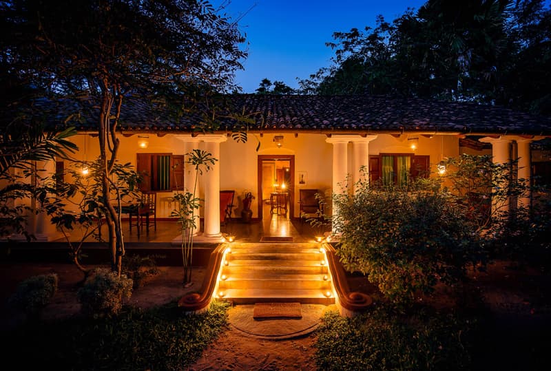 The white-columned walawwa veranda at deep-blue dusk, every step of the entrance stair lined with glowing candle lanterns, warm lamplight under the clay-tile roof