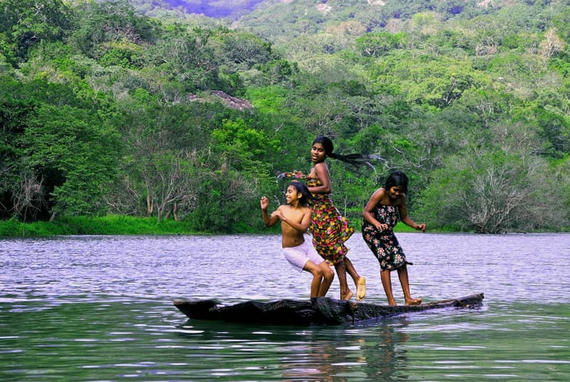 Three village children laughing and balancing on a wooden dugout canoe on the lake, jungle hillside behind