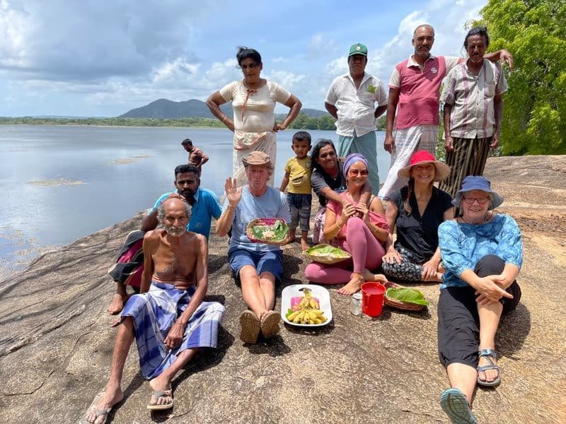 A group of villagers and retreat guests sitting on a large lakeside rock, sharing a picnic of bananas and food wrapped in banana leaves.