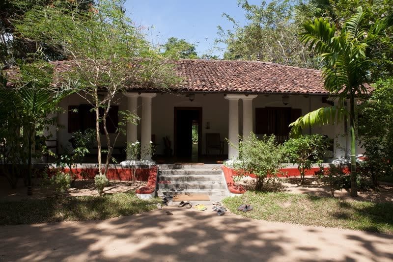 White colonial-era manor house with red tile roof and columned veranda, pairs of sandals left at the foot of its steps, surrounded by tropical trees.